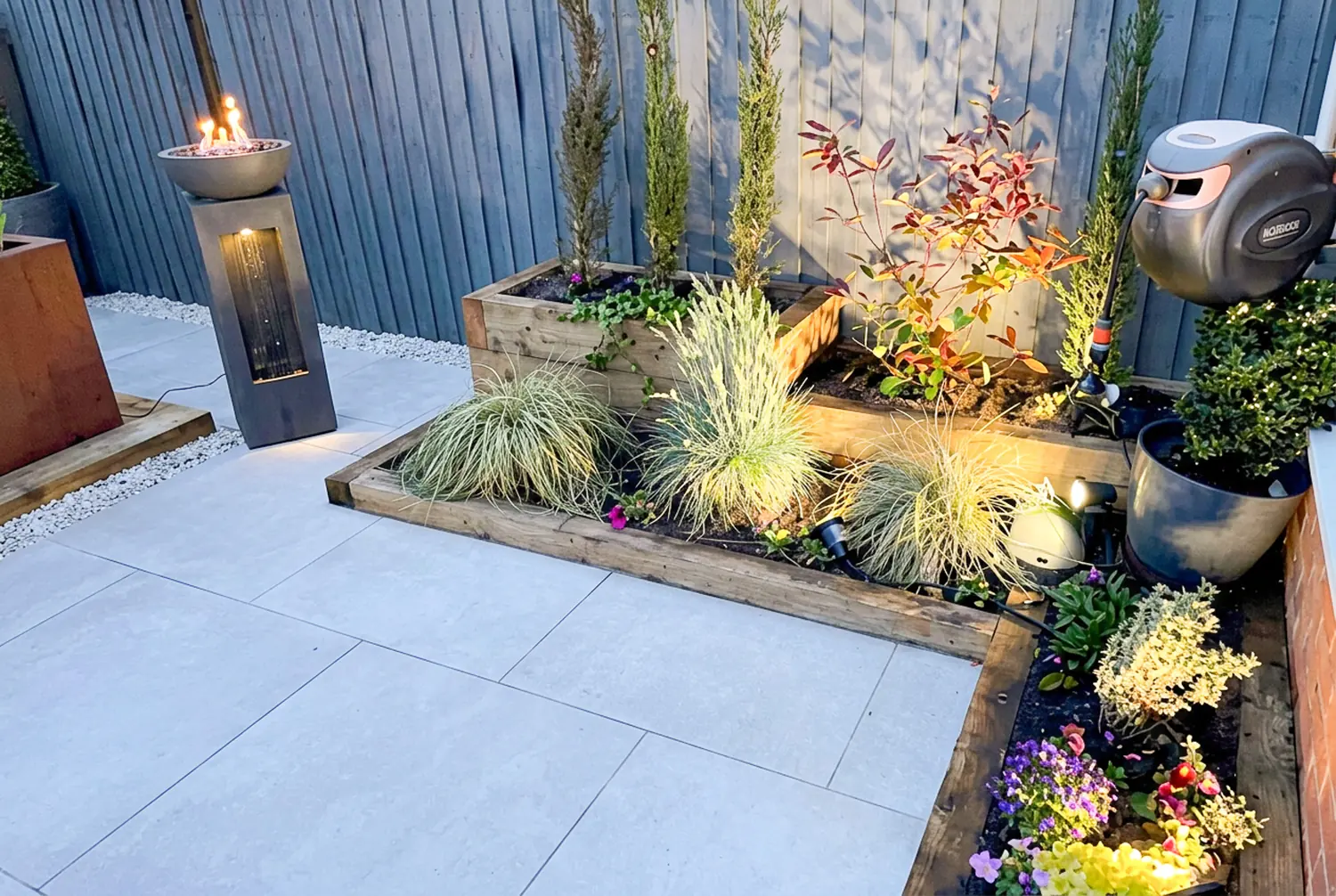 Porcelain paving beside timber sleeper raised beds with ornamental grasses and garden uplighters