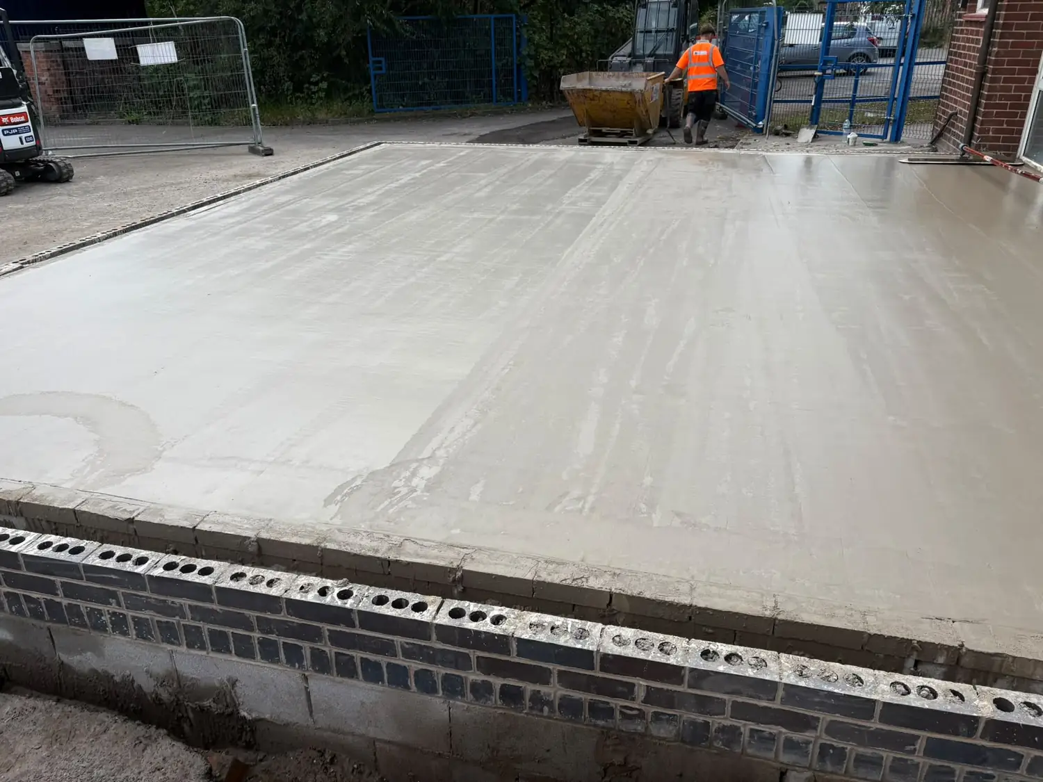 Freshly poured concrete slab being levelled off, with a groundworker and skip behind